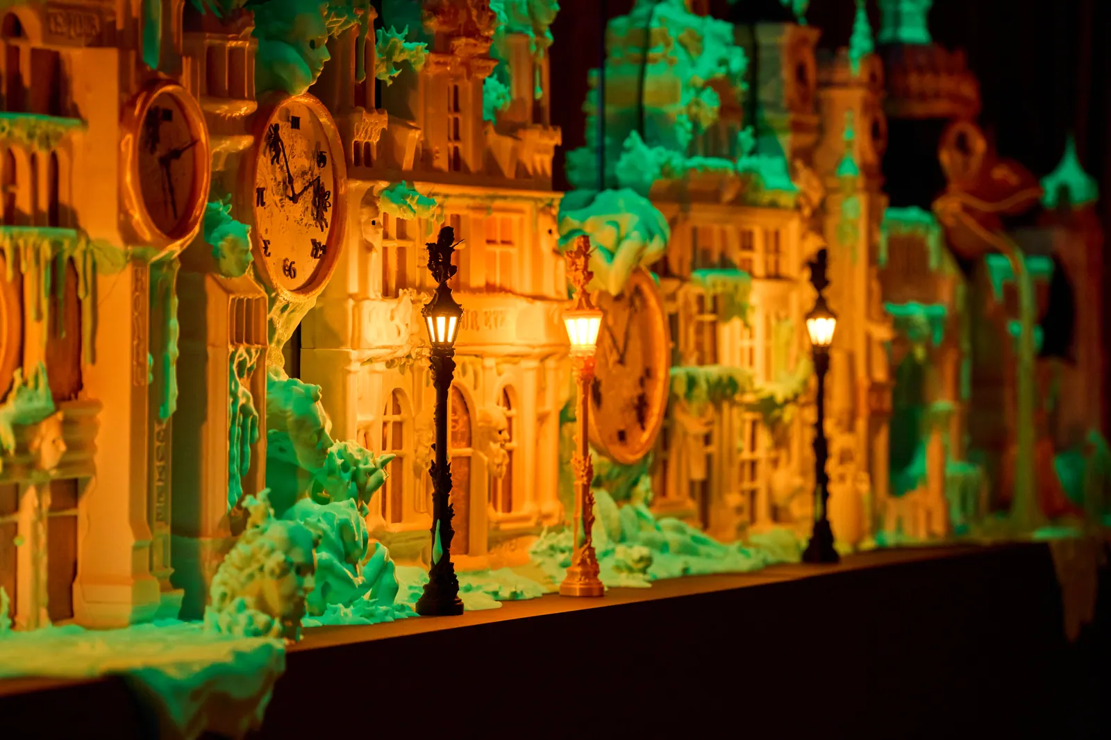 A brightly lit miniature fantasy city and green sculptural objects displayed in a dark gallery