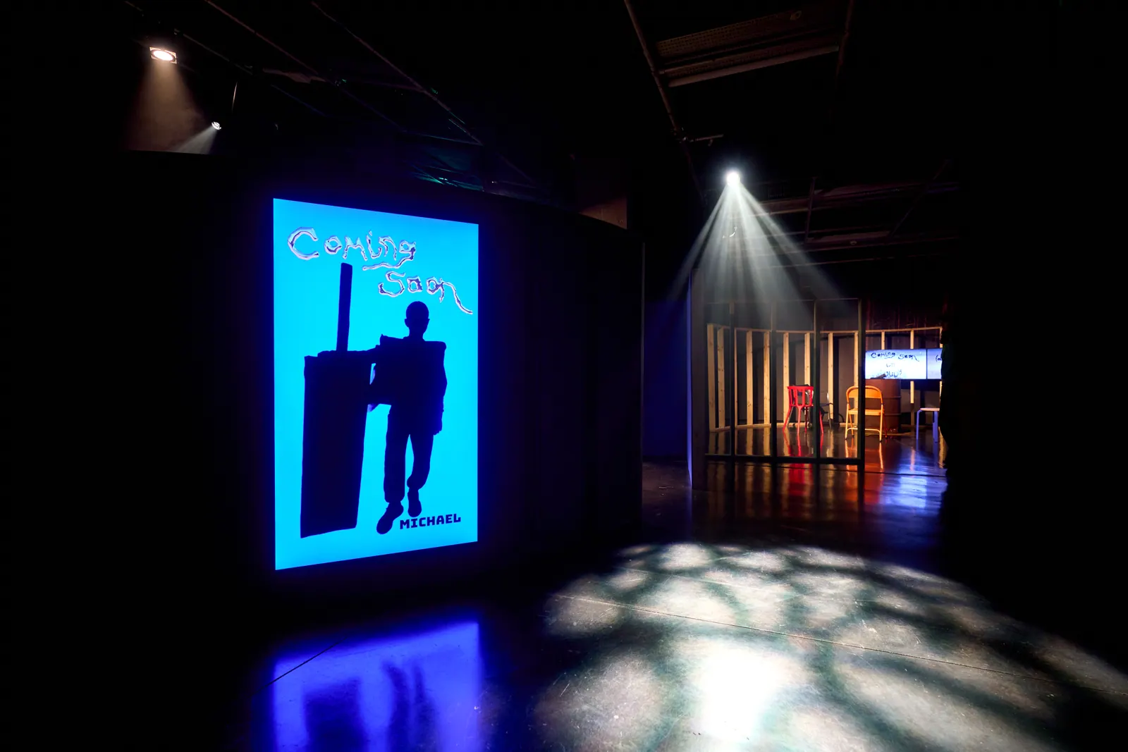 A dark gallery with a bright blue screen, a silhouetted figure and the wooden installation beyond